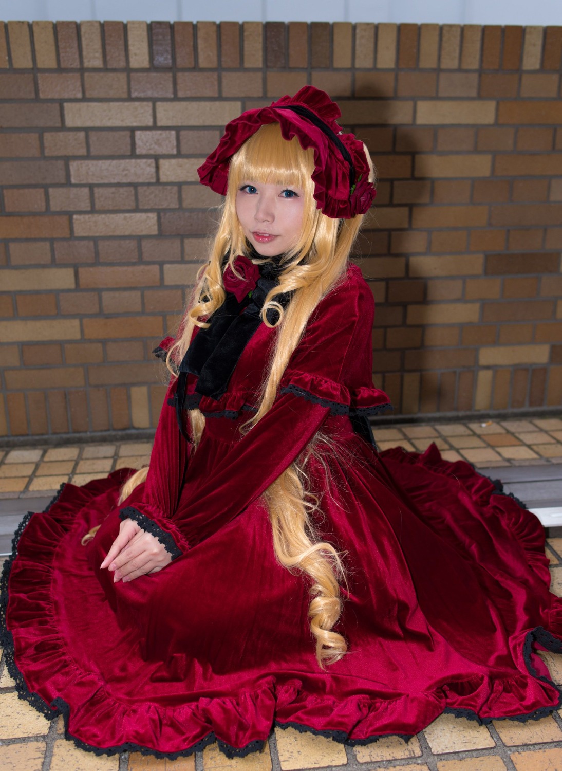 1girl blonde_hair blue_eyes bonnet brick_wall capelet dress drill_hair flower frills lolita_fashion long_hair long_sleeves looking_at_viewer red_capelet red_dress rose shinku sitting solo tile_floor tiles