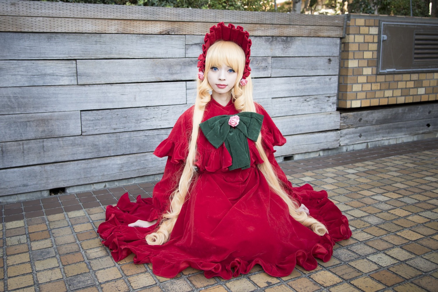 1girl blonde_hair blue_eyes bonnet bow brick_wall capelet dress fence flower looking_at_viewer on_floor pavement red_dress shinku sitting smile solo stone_floor tile_floor tiles wall