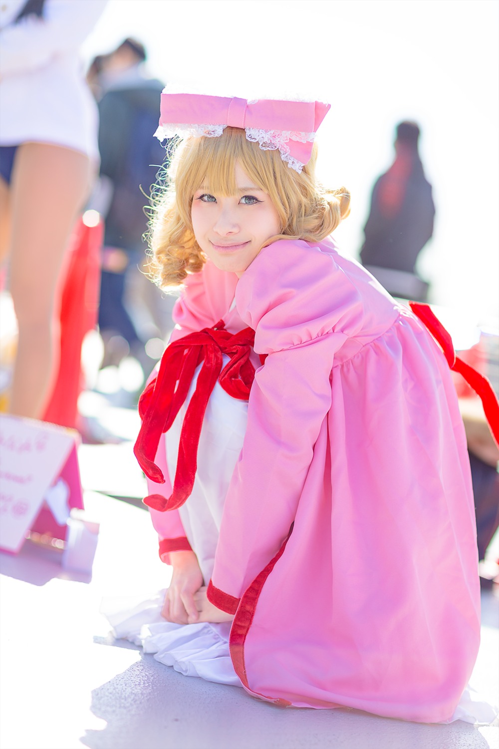 1girl 3d blonde_hair blue_eyes blurry blurry_background blurry_foreground book_stack bow depth_of_field dress drill_hair frills hinaichigo looking_at_viewer motion_blur photo pink_bow pink_dress ribbon sitting smile solo solo_focus