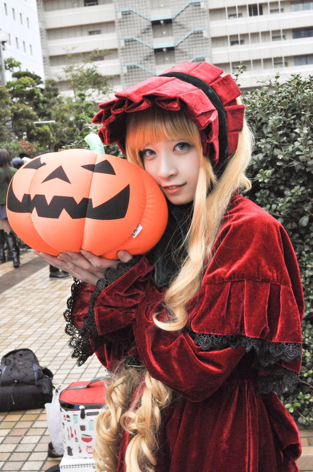 1girl blonde_hair blue_eyes bonnet building chain-link_fence city day dress fence hat jack-o'-lantern lips lolita_fashion long_hair long_sleeves looking_at_viewer outdoors pumpkin red_dress shinku solo traditional_media tree