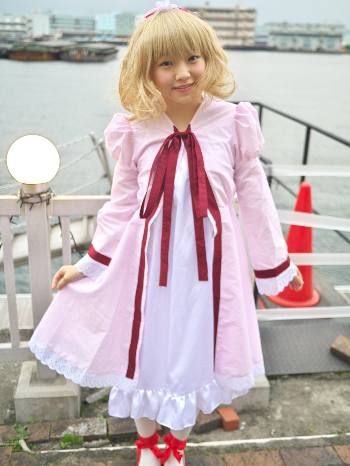 1girl alice_margatroid blonde_hair blue_eyes blurry building city day depth_of_field dress hairband hinaichigo long_sleeves looking_at_viewer outdoors railing realistic red_ribbon ribbon short_hair sky smile solo standing