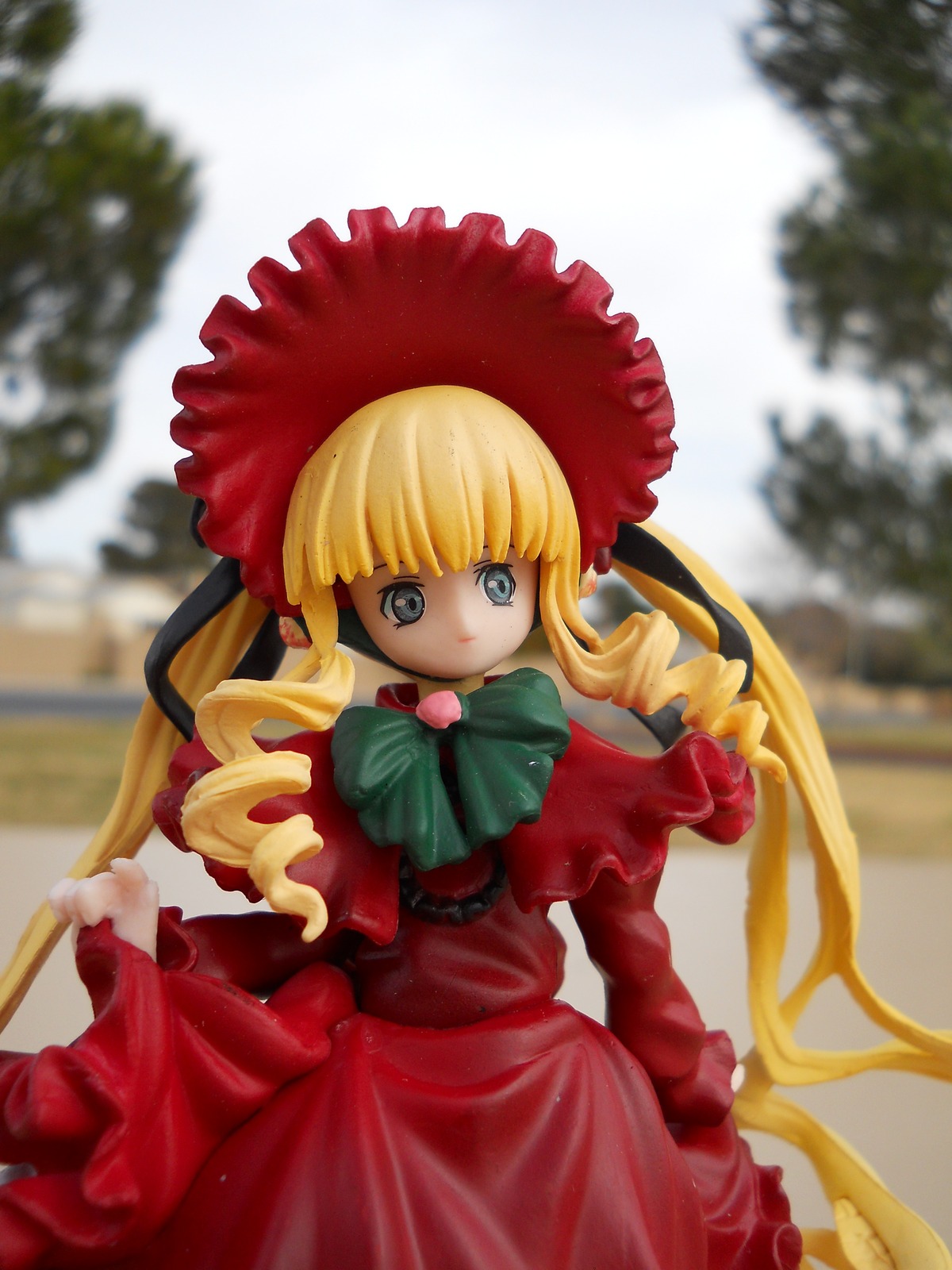 1girl bangs blonde_hair blue_eyes blurry blurry_background blurry_foreground bonnet bow bowtie capelet depth_of_field doll dress drill_hair flower frills green_bow long_hair long_sleeves looking_at_viewer outdoors photo red_dress shinku sidelocks solo standing twin_drills twintails