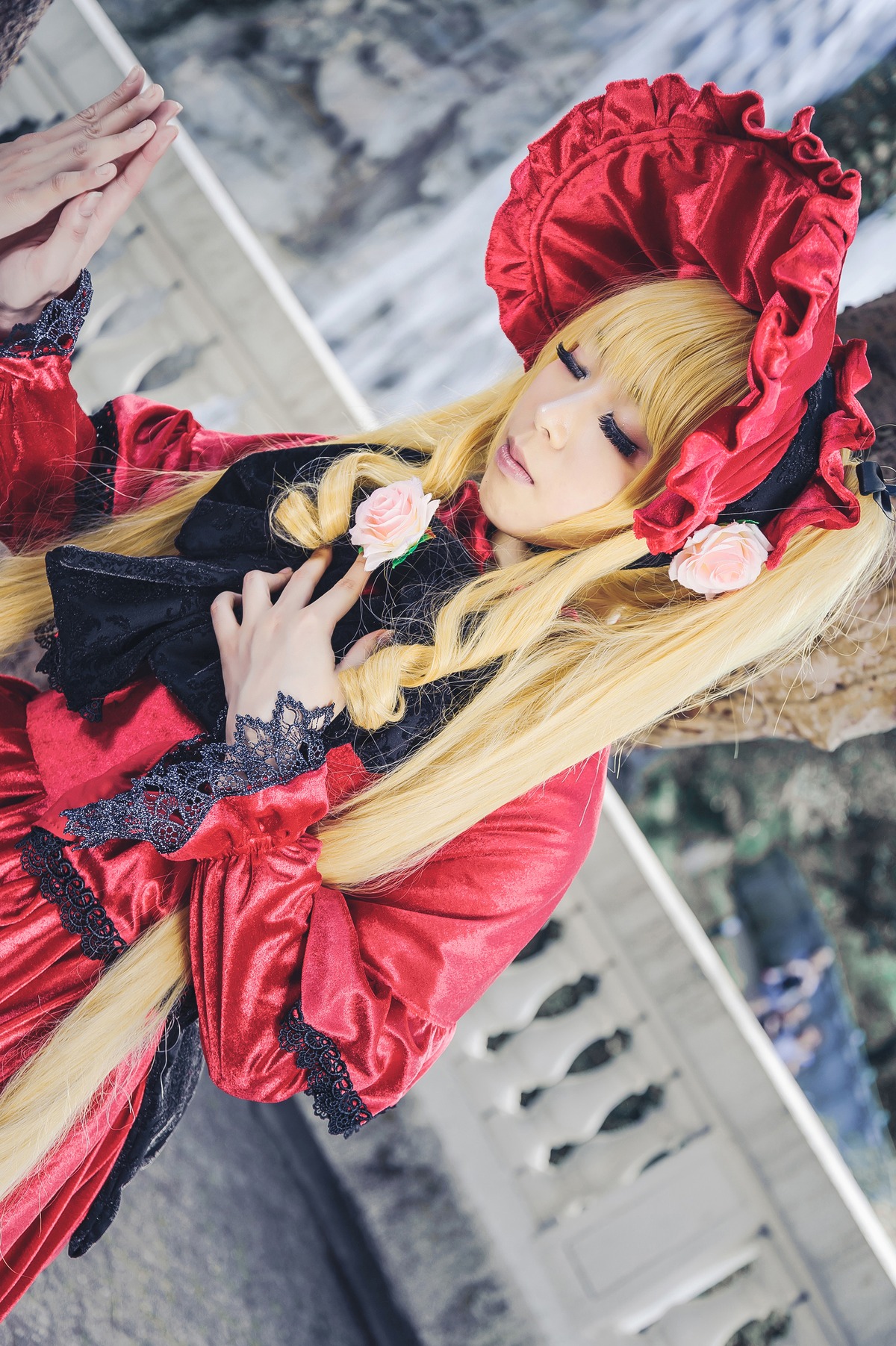 1girl blonde_hair blurry blurry_background blurry_foreground bonnet closed_eyes depth_of_field dress flower hat long_hair long_sleeves outdoors photo red_dress rose shinku solo solo_focus