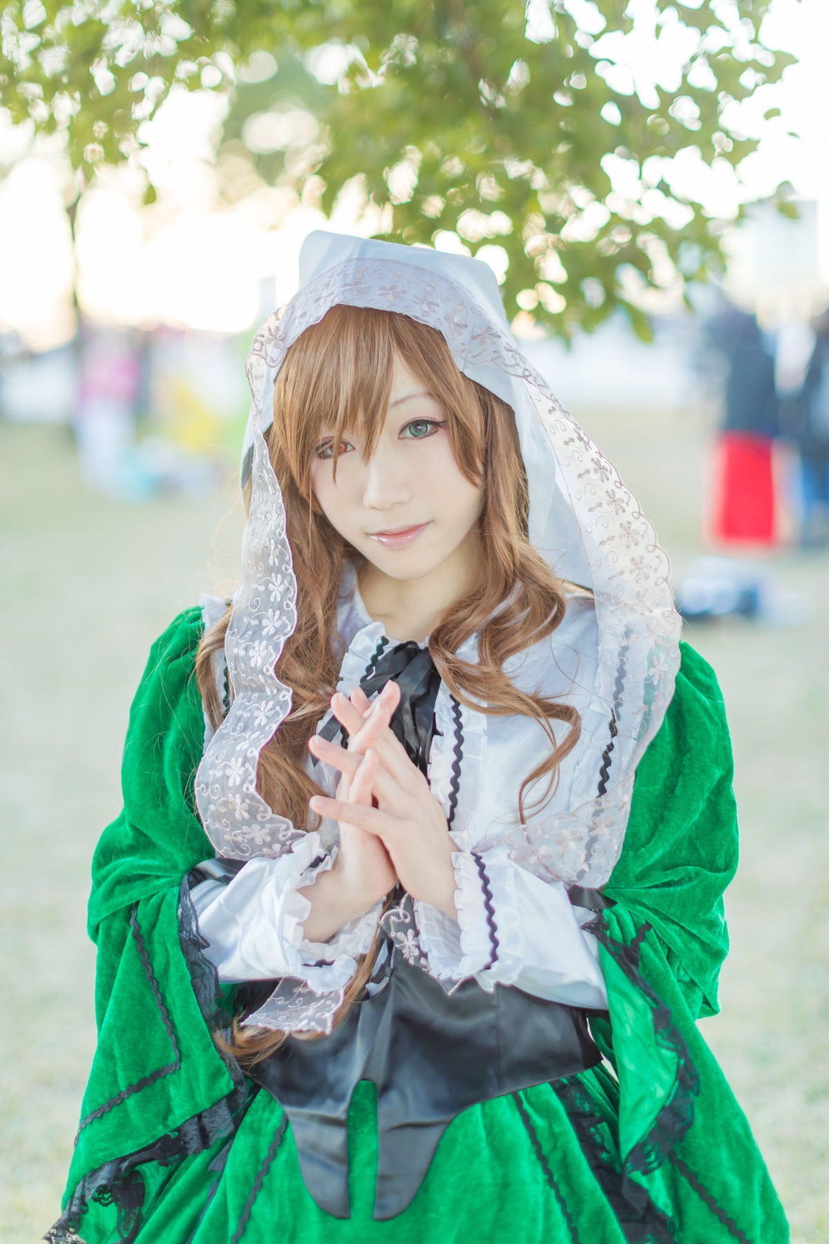 1girl blonde_hair blurry blurry_background blurry_foreground brown_hair depth_of_field dress flower green_dress green_eyes hands_clasped hands_together hood interlocked_fingers lips long_hair long_sleeves looking_at_viewer own_hands_together ribbon smile solo suiseiseki