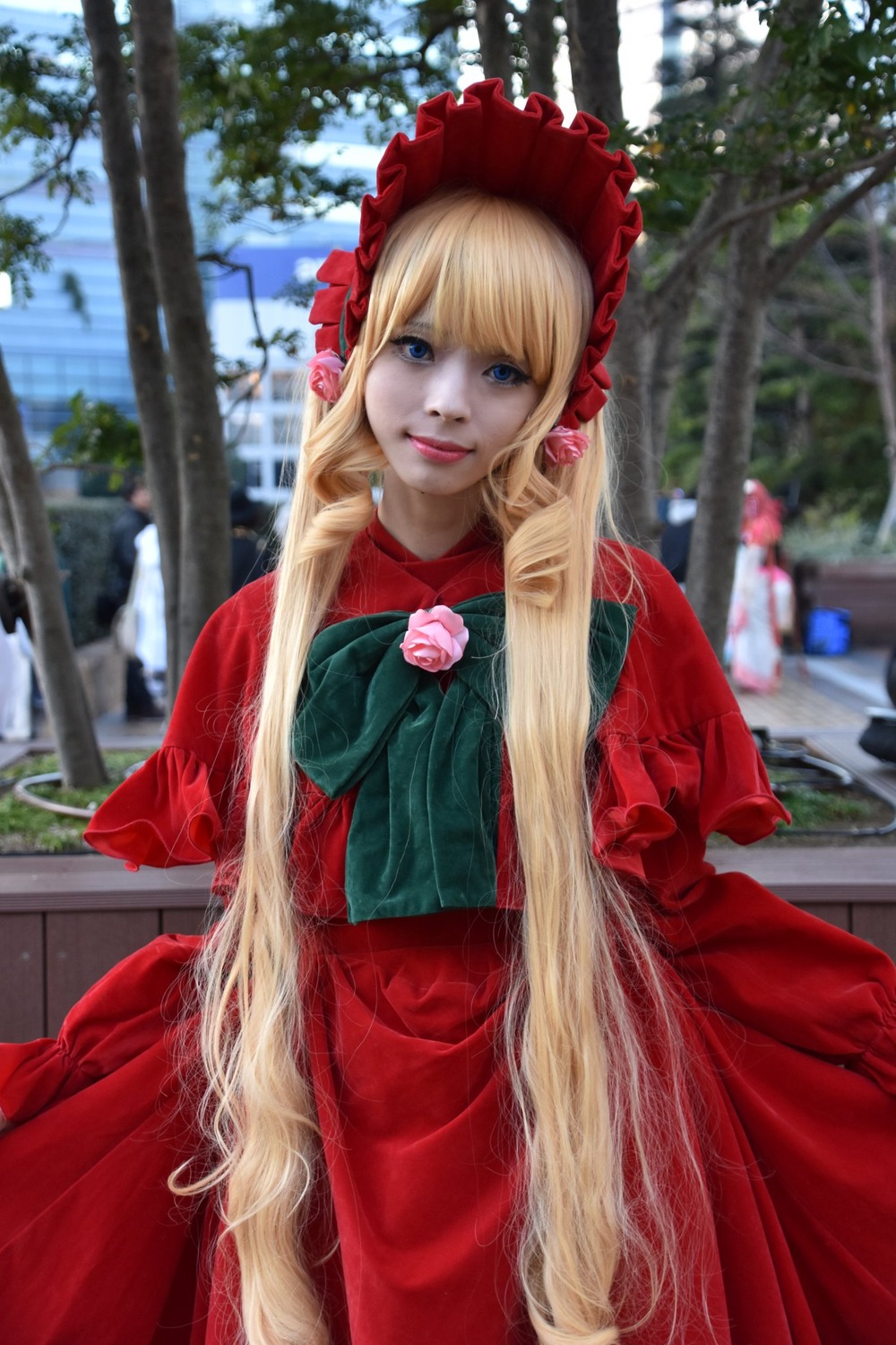 1girl blonde_hair blue_eyes blurry blurry_background bonnet building capelet depth_of_field dress flower lips long_hair long_sleeves looking_at_viewer photo red_capelet red_dress rose shinku smile solo very_long_hair