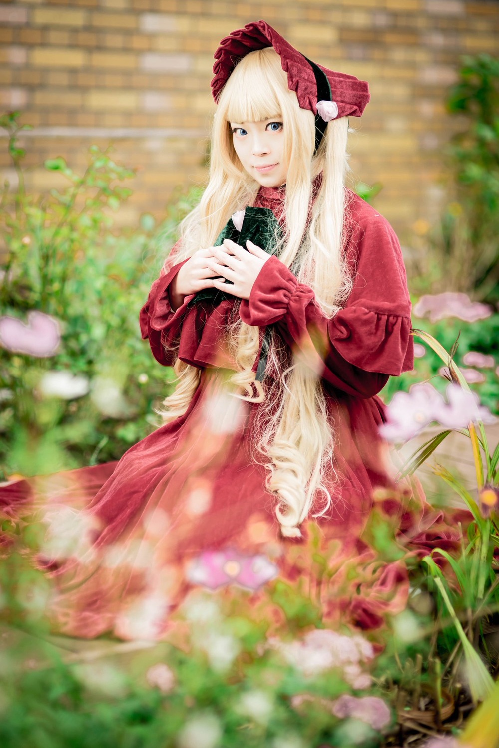 1girl bangs blonde_hair blue_eyes blurry blurry_background blurry_foreground bonnet depth_of_field dress flower long_hair long_sleeves looking_at_viewer outdoors red_dress shinku smile solo standing