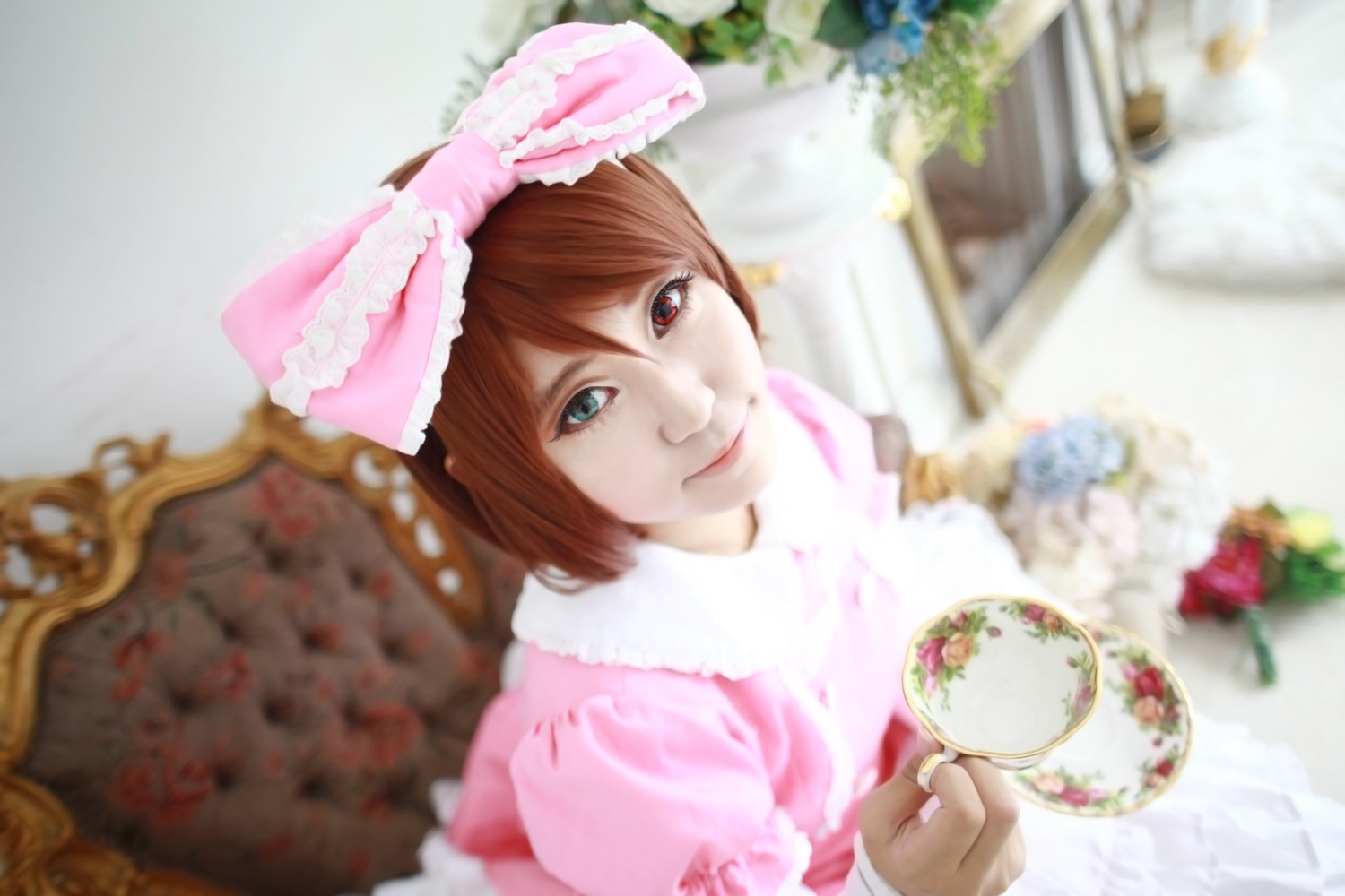 1girl 3d bangs blurry blurry_background blurry_foreground bow brown_hair cup depth_of_field dress food frills heterochromia lips looking_at_viewer photo pink_dress smile solo souseiseki