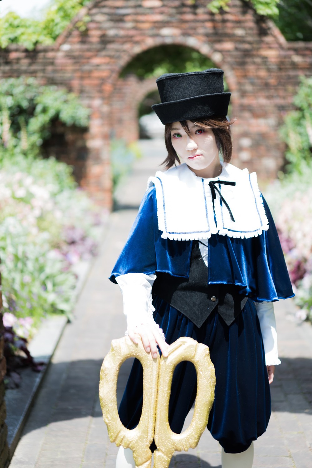 1girl blue_dress blurry blurry_background blurry_foreground brown_hair capelet depth_of_field dress flower green_eyes hat heterochromia long_sleeves looking_at_viewer outdoors photo_background short_hair solo souseiseki standing