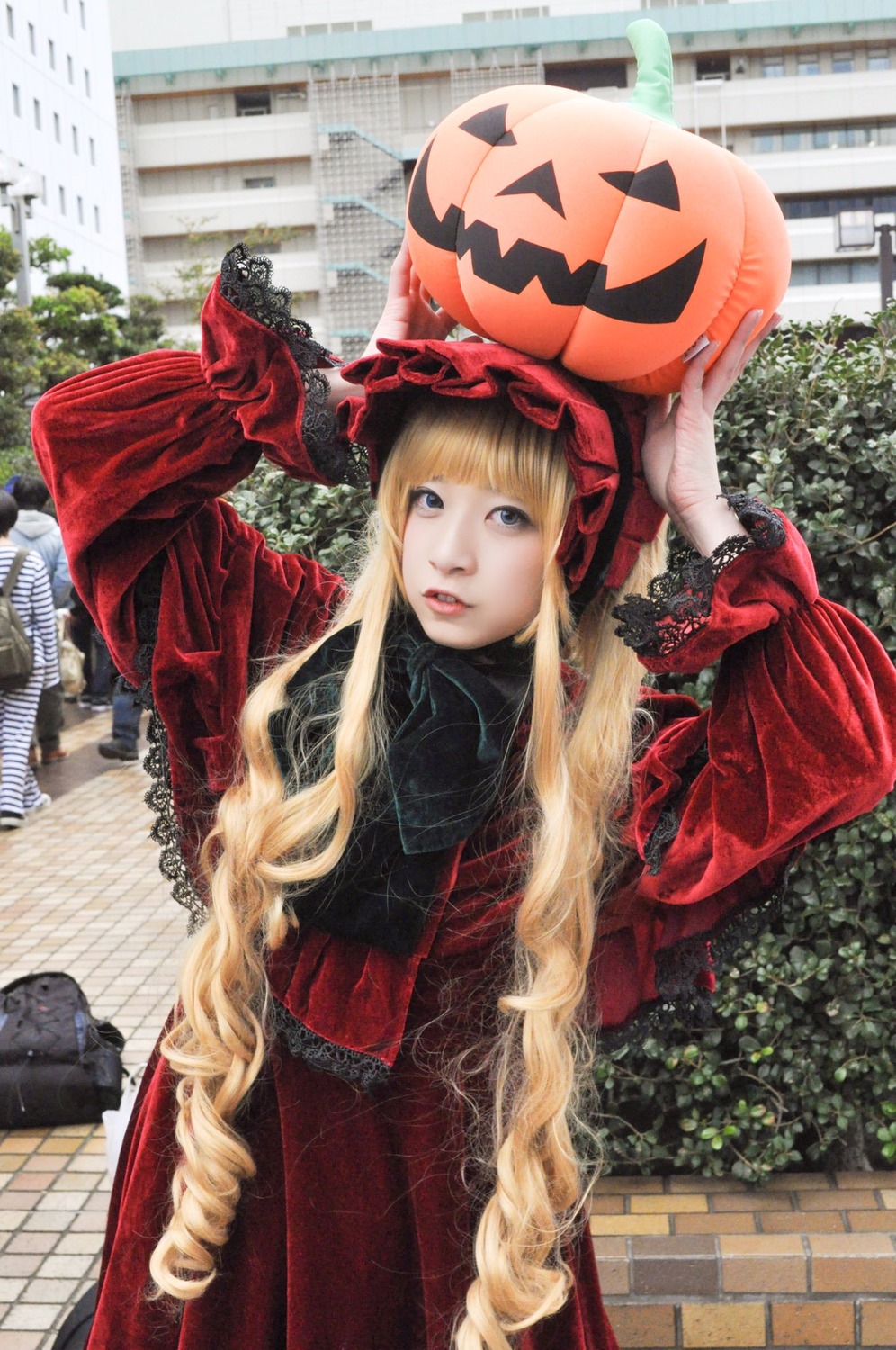 1girl arms_up bangs blonde_hair brick_wall building chain-link_fence city day dress drill_hair fence hat jack-o'-lantern lips long_hair long_sleeves looking_at_viewer outdoors pumpkin red_dress shinku solo street very_long_hair