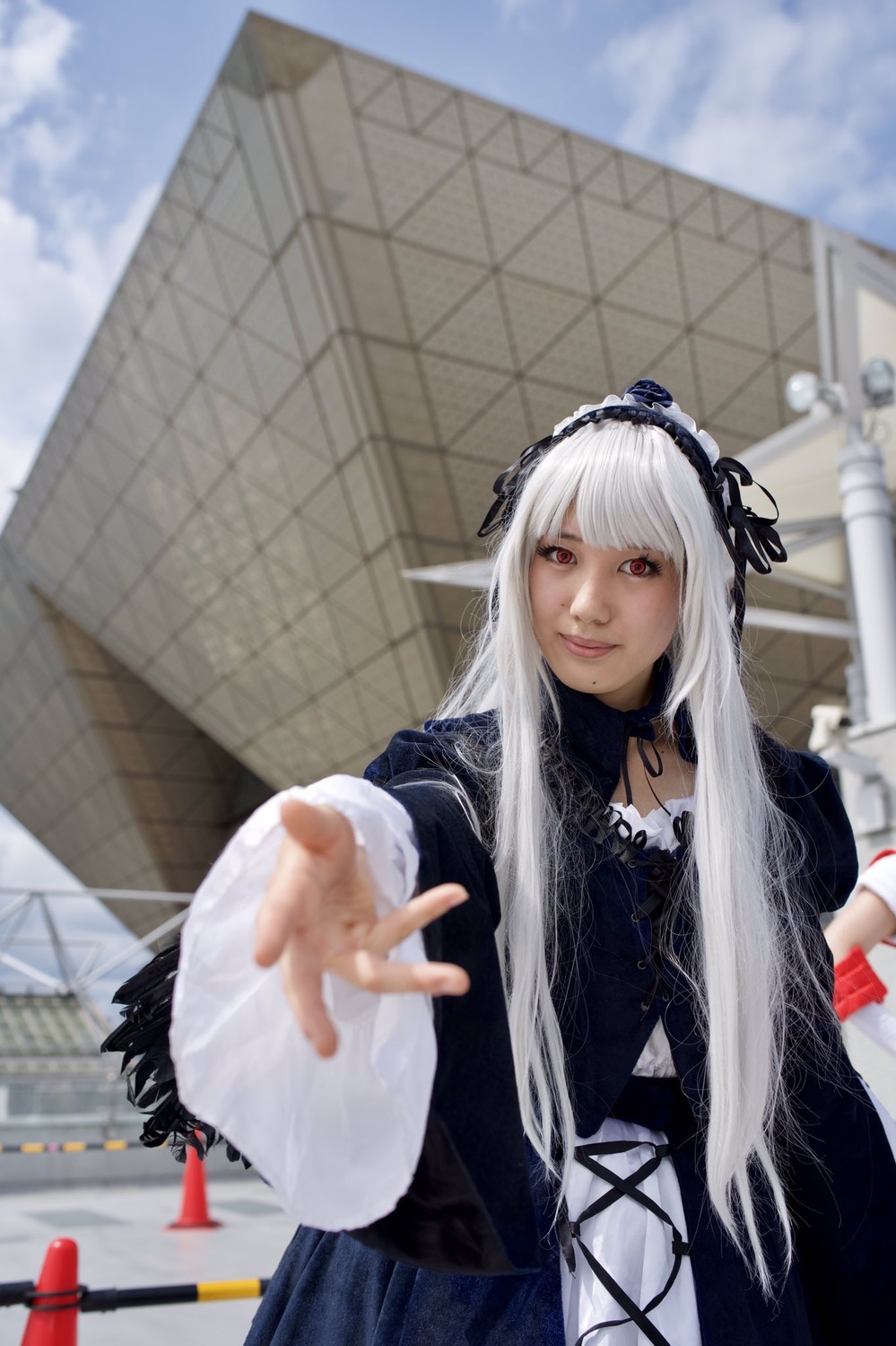 1girl 3d blurry blurry_background blurry_foreground building city day depth_of_field dress gothic_lolita hairband lolita_fashion long_hair long_sleeves looking_at_viewer outstretched_arm outstretched_hand photo red_eyes solo suigintou wings