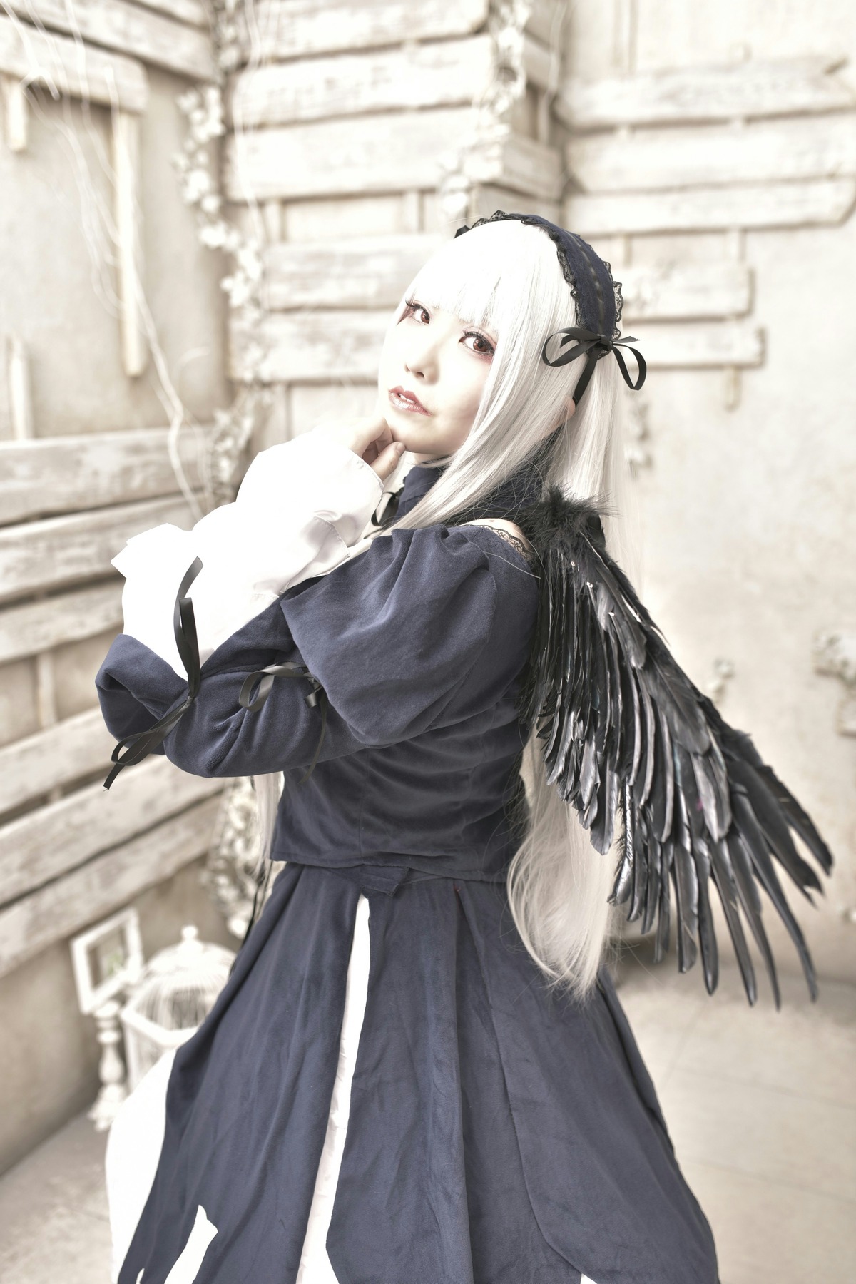 1girl bangs black_dress black_ribbon black_wings blurry blurry_background blurry_foreground building church depth_of_field dress feathered_wings feathers hairband long_hair long_sleeves looking_at_viewer outdoors photo ribbon solo standing suigintou white_hair wings