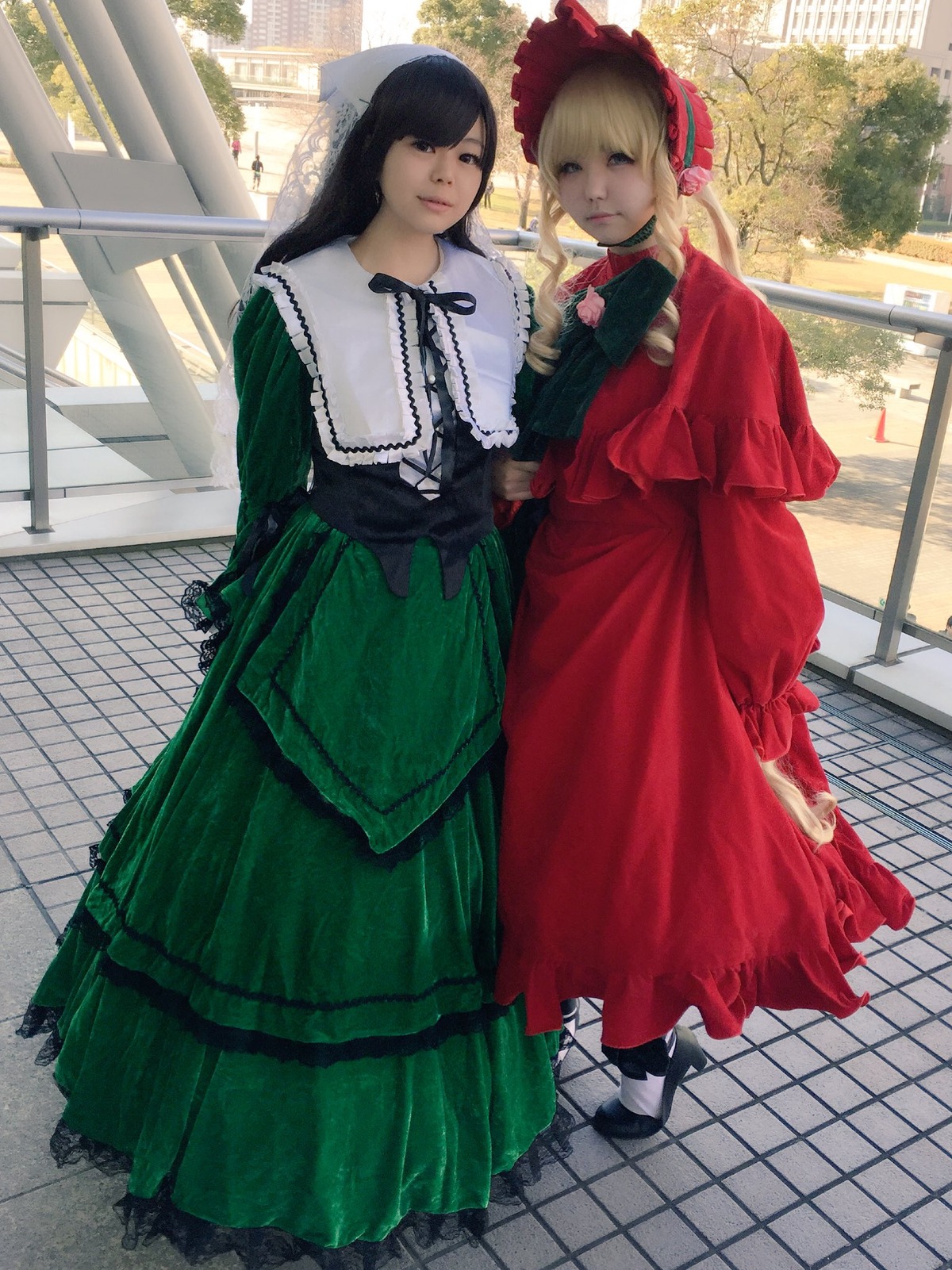 2girls bangs black_hair blonde_hair bonnet building bush capelet chain-link_fence day dress fence green_dress long_hair long_skirt long_sleeves looking_at_viewer multiple_cosplay multiple_girls outdoors pavement red_dress shoes standing tagme tile_floor tiles white_legwear window