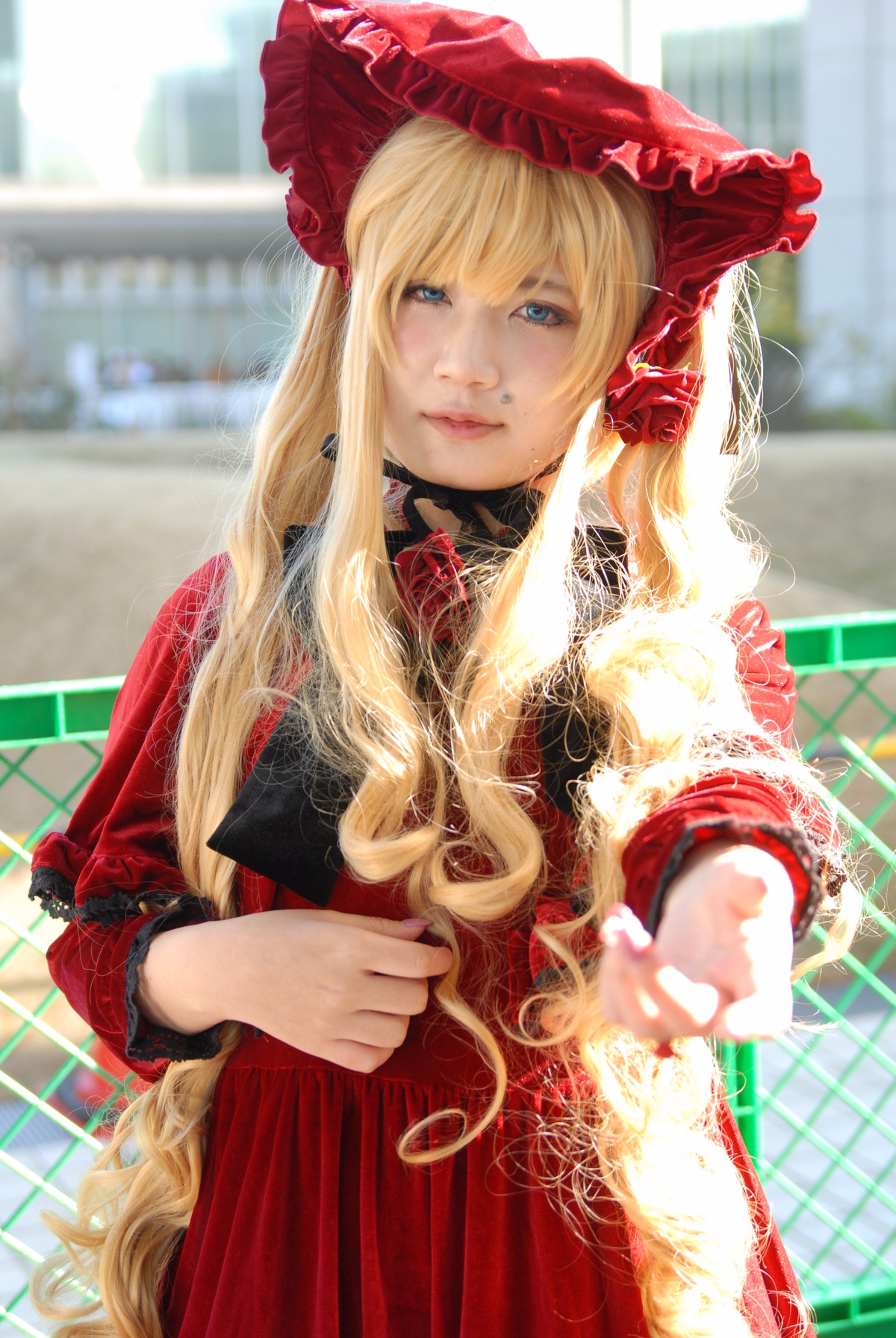 1girl blonde_hair blue_eyes blurry chain-link_fence depth_of_field dress fence flower lips long_hair long_sleeves looking_at_viewer red_dress shinku smile solo