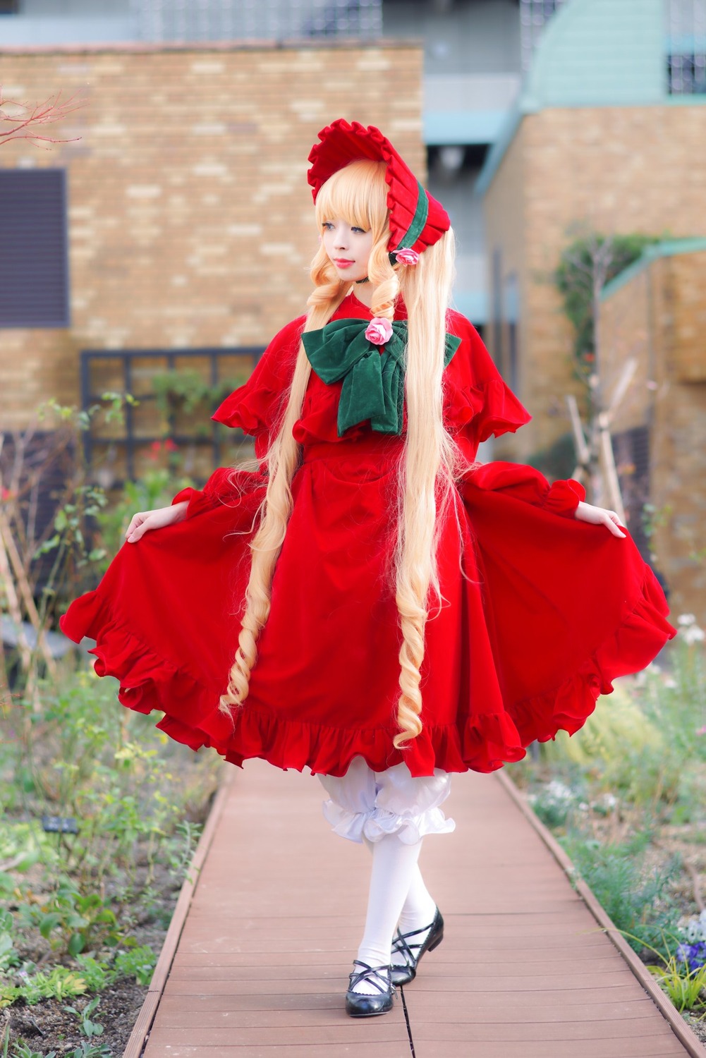 1girl blonde_hair blurry blurry_background bonnet bow capelet depth_of_field dress flower green_eyes long_hair outdoors pantyhose photo red_capelet red_dress shinku shoes solo standing white_legwear
